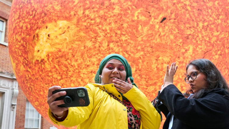 Two young women smiling as they take selfies in front of Luke Jerram's Helios